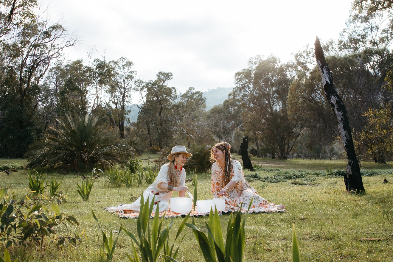 Two women sharing crystal singing bowls in a grassy landscape