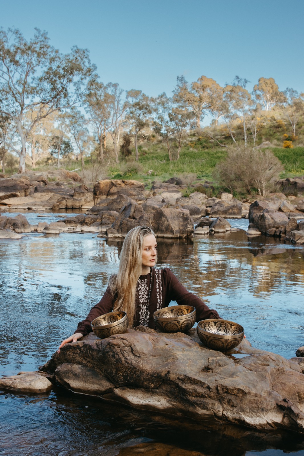 Shana-lea seated on river rocks with three singing bowls, water and gum trees behind her